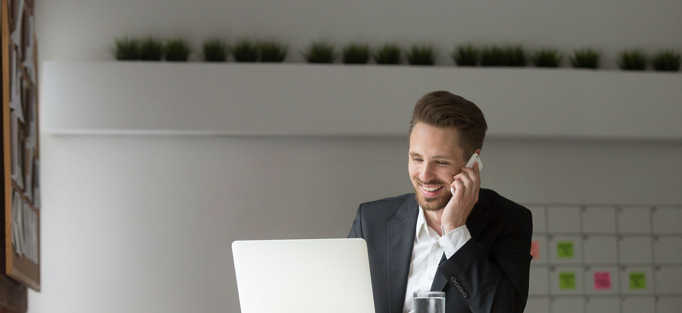 Smiling businessman wearing suit sitting at working desk using typing on laptop, making answering call, talking on the phone, consulting client about email by cell, holding mobile interview in office