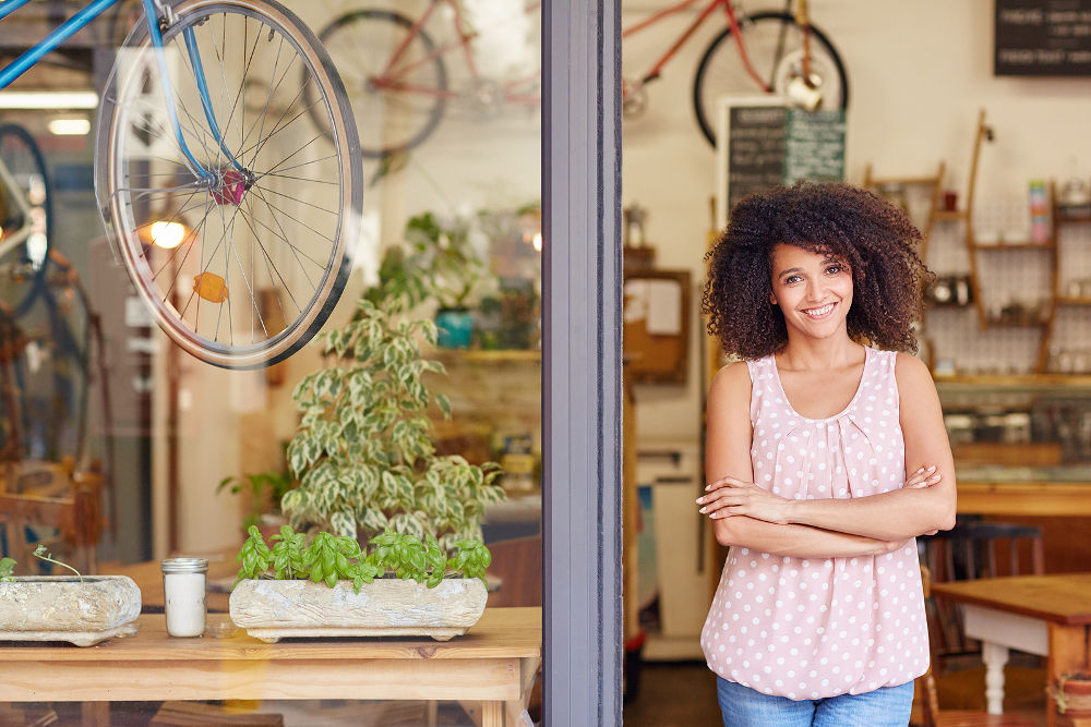 Young mixed race woman smiling, while standing in the door of her cafe with her arms folded proud to be the owner of a small business