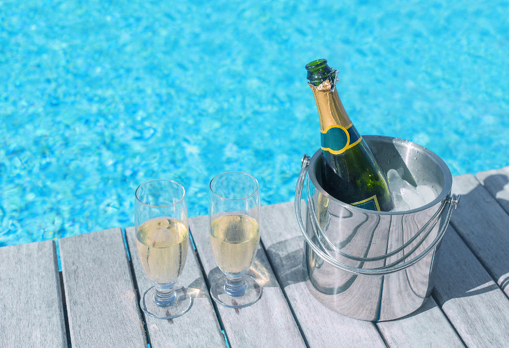 Cold champagne bottle in ice bucket and two glasses of champagne on the deck by the swimming pool