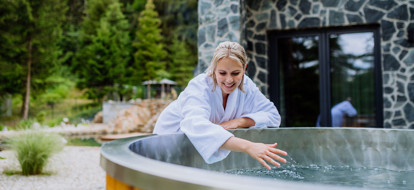 Young woman in bathrobe, checking temperature, ready for home spa procedure in hot tub outdoors. Wellness, body care, hygiene concept.