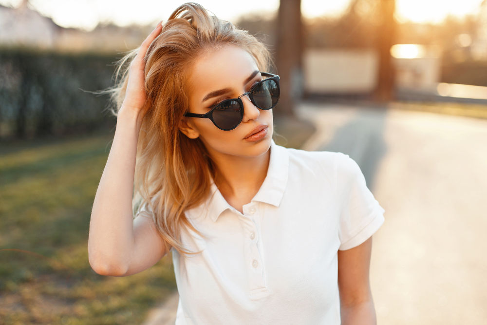 Beautiful young woman wearing sunglasses and white polo shirt walking at sunset