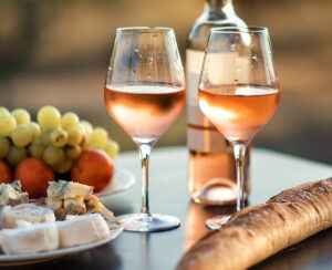 Bottle of rose wine and two full glasses of wine on table in heart of Provence, France with french bread, cheese, ham, grapes and peaches with olive trees on background in sunset. Travel in Provence.