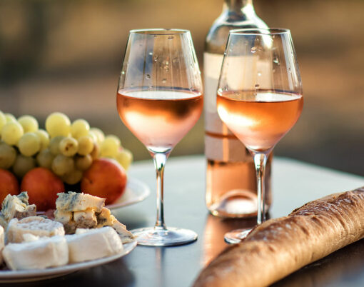 Bottle of rose wine and two full glasses of wine on table in heart of Provence, France with french bread, cheese, ham, grapes and peaches with olive trees on background in sunset. Travel in Provence.