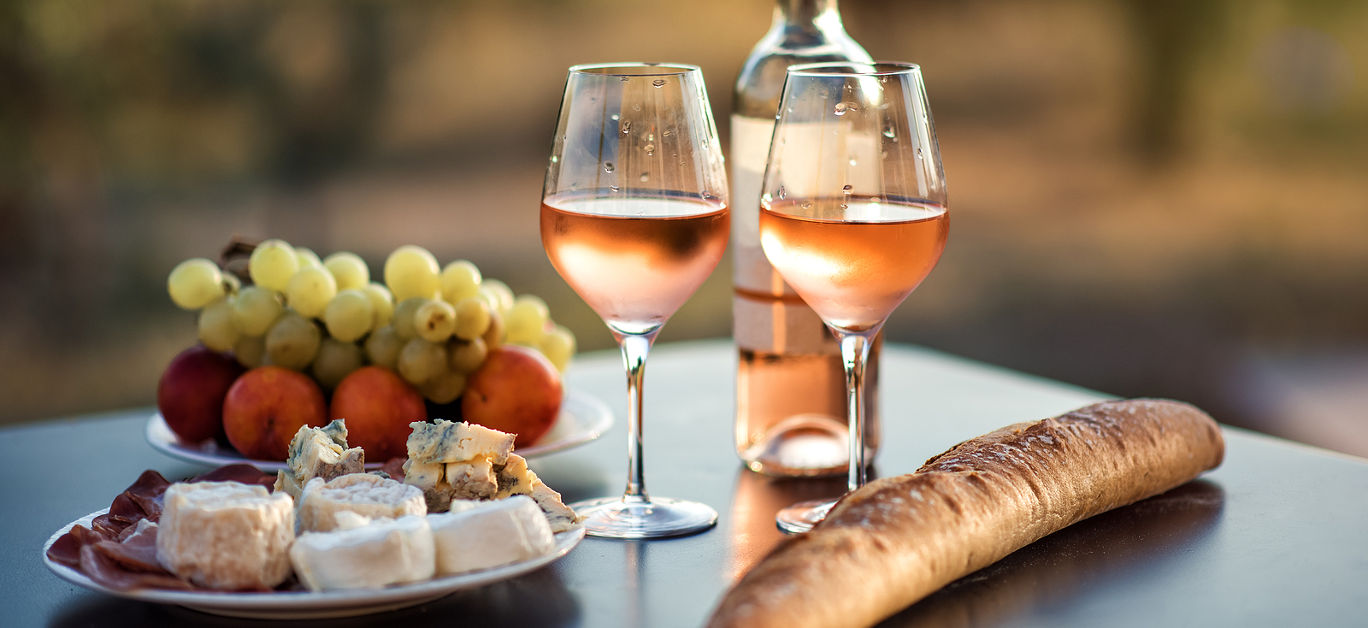 Bottle of rose wine and two full glasses of wine on table in heart of Provence, France with french bread, cheese, ham, grapes and peaches with olive trees on background in sunset. Travel in Provence.