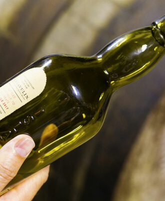 Someone holds a bottle of 16 years aged Lagavulin single malt whisky at the Lagavulin whisky distillery with old oak cask on the background.