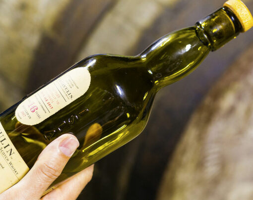 Someone holds a bottle of 16 years aged Lagavulin single malt whisky at the Lagavulin whisky distillery with old oak cask on the background.