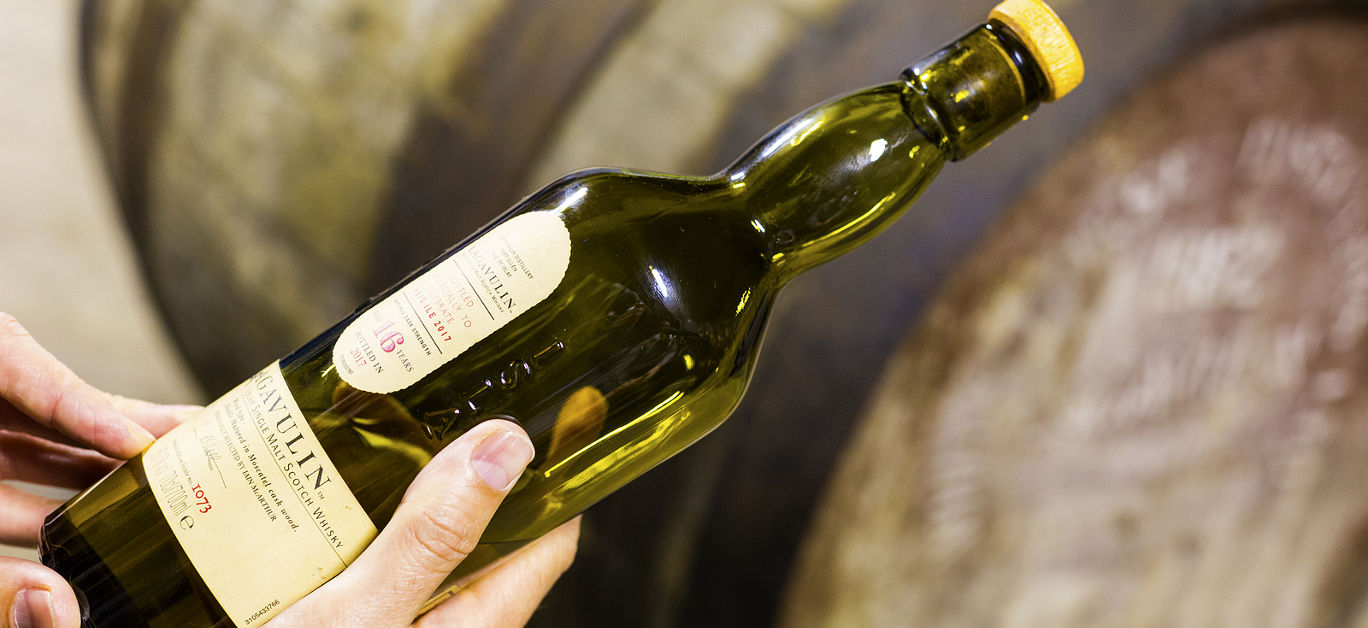 Someone holds a bottle of 16 years aged Lagavulin single malt whisky at the Lagavulin whisky distillery with old oak cask on the background.