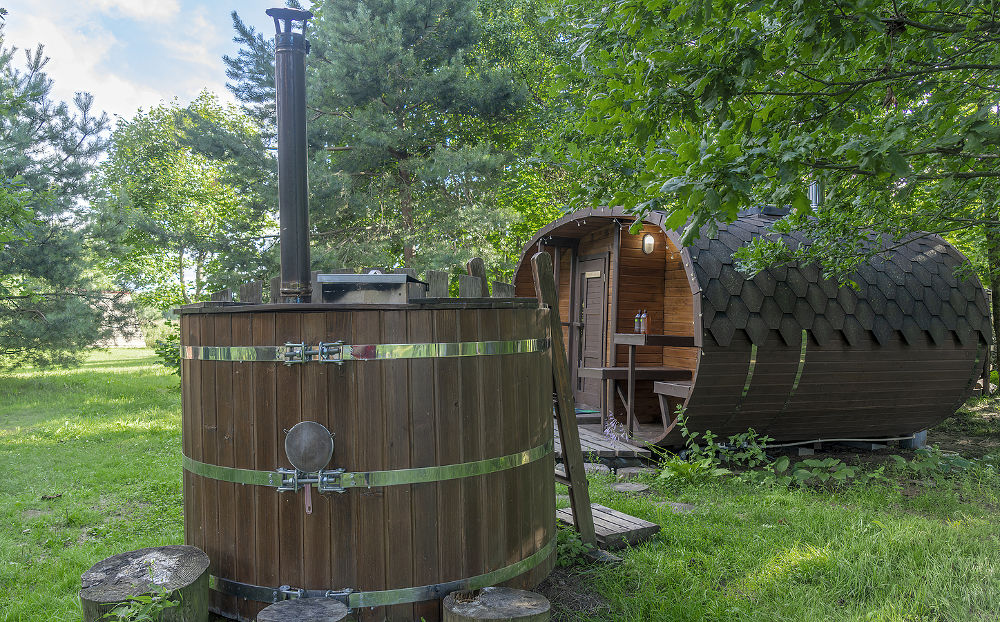 Outdoor wooden barrel sauna in the garden. In foreground wooden bathtub with fireplace.