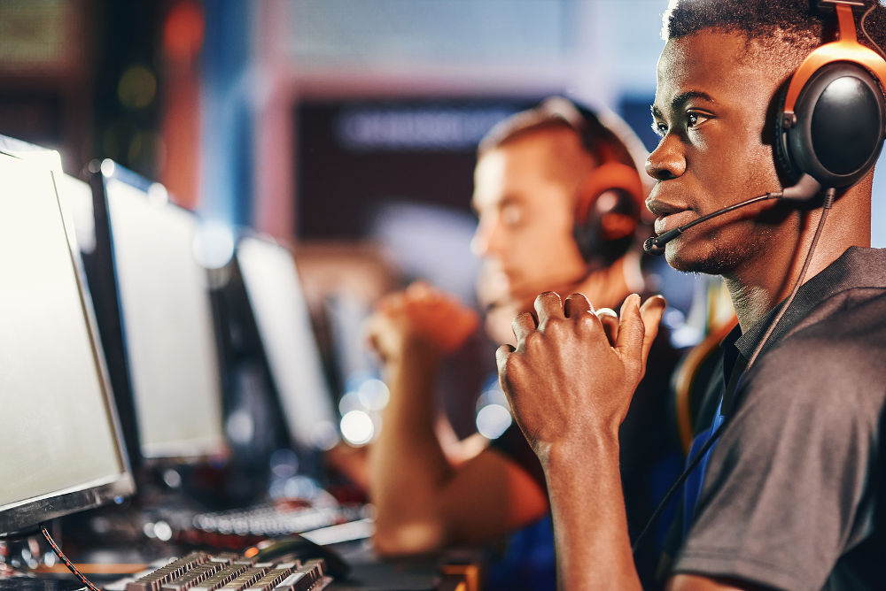 Side view of a fully concentrated african guy, professional cybersport gamer looking at PC screen while sitting in gaming club or internet cafe and participating eSport tournament