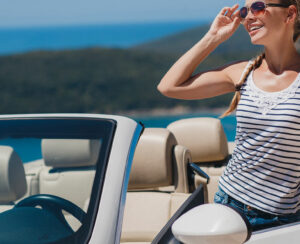 Portrait of a beautiful young woman,brunette with a long braid,wearing dark glasses,a striped t-shirt,posing alone in a white convertible car on a blue sky background,mountains and blue sea