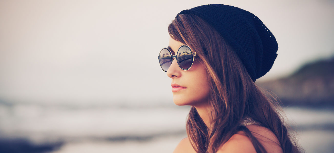 Fashion portrait of young hipster woman with hat and sunglasses on the beach at sunset, retro style color tones