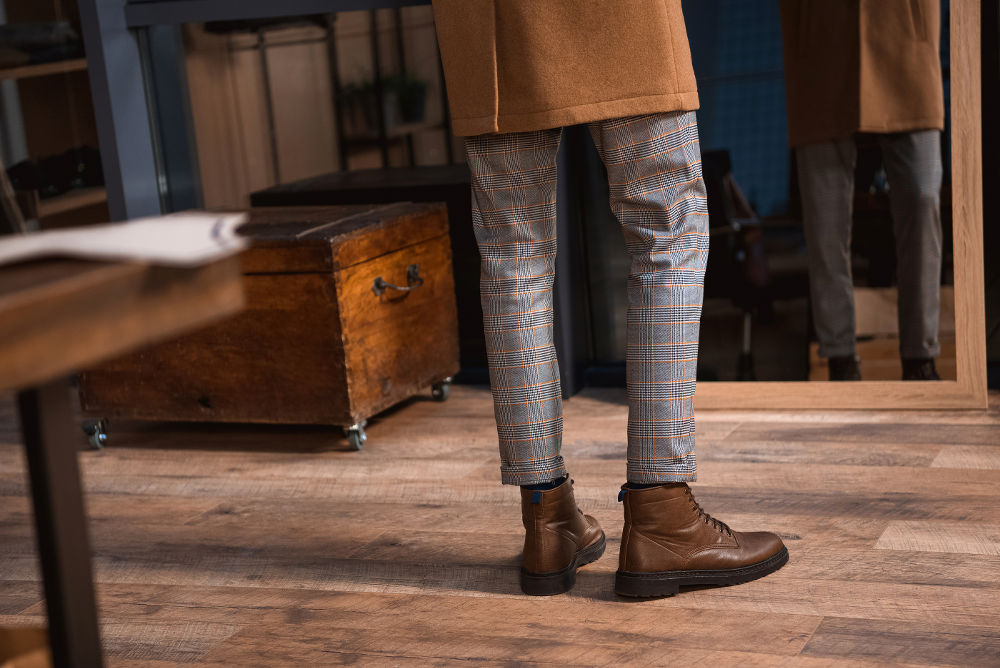 low section of man in leather boots and overcoat standing near mirror in boutique