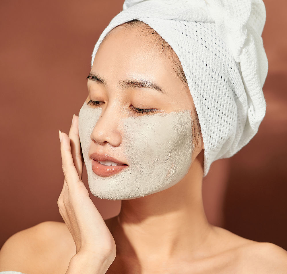 Beautiful woman with clay facial mask over orange background. Beauty treatment and spa concept.