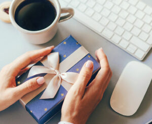 Blue gift box decorated with a golden ribbon bow in female hands against the background of the office workplace, gift delivery, coffee break during office hours, corporate greetings and parties.