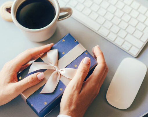 Blue gift box decorated with a golden ribbon bow in female hands against the background of the office workplace, gift delivery, coffee break during office hours, corporate greetings and parties.