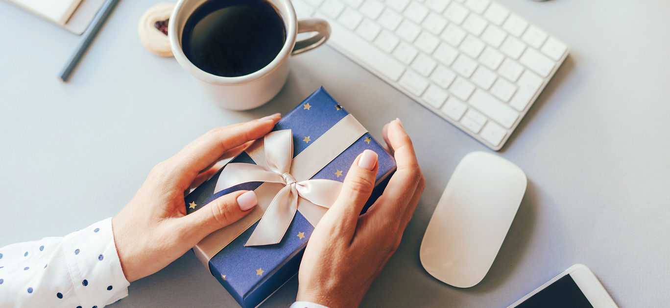 Blue gift box decorated with a golden ribbon bow in female hands against the background of the office workplace, gift delivery, coffee break during office hours, corporate greetings and parties.