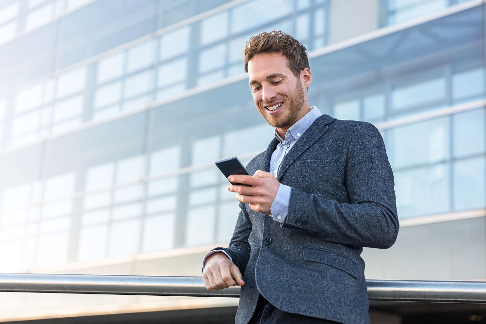 Businessman using mobile phone app texting outside of office in urban city with skyscrapers buildings in the background