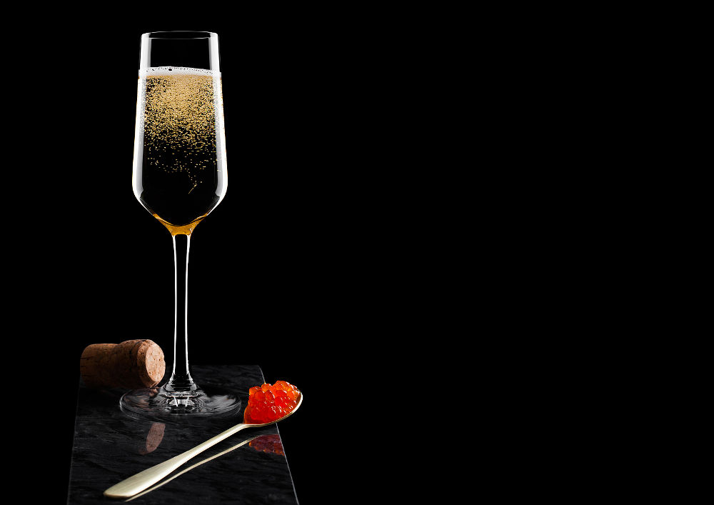 Elegant glass of yellow champagne with red caviar on golden spoon and cork of caviar on marble board on black.