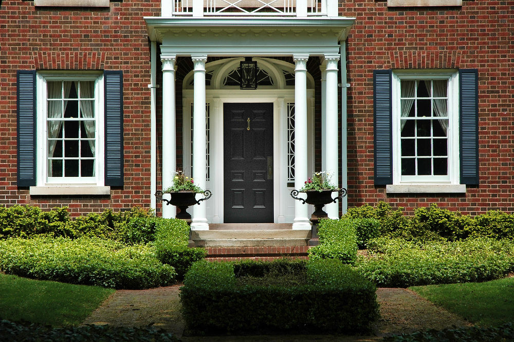 Red brick house with black shutters and green shrubs with front door