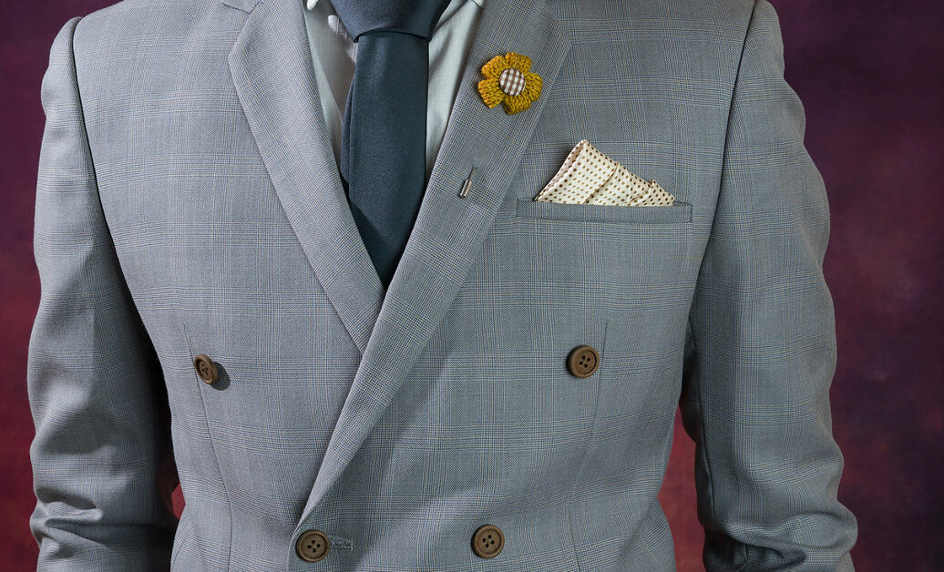 Man in double breasted grey suit plaid texture blue necktie flower brooch and dot pattern handkerchief close up