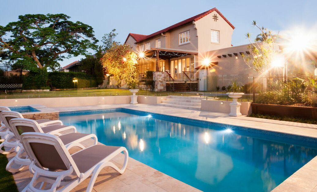 Luxury house and swimming pool decoration at an evening with flashing lights around the garden with trees there is a green lawn around the waterside with chairs