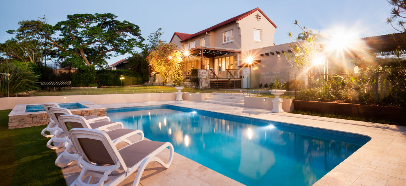 Luxury house and swimming pool decoration at an evening with flashing lights around the garden with trees there is a green lawn around the waterside with chairs