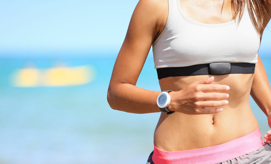 Runner woman with heart rate monitor running on beach with watch and sports bra top. Beautiful fit female fitness model training and working out outside in summer at part of healthy lifestyle.