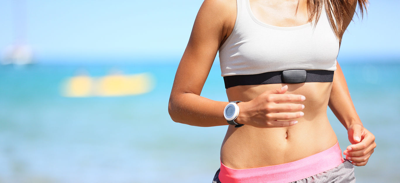 Runner woman with heart rate monitor running on beach with watch and sports bra top. Beautiful fit female fitness model training and working out outside in summer at part of healthy lifestyle.